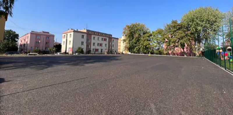 Widok na nowo wyasfaltowany plac z budynkami w tle oraz drzewami po bokach.