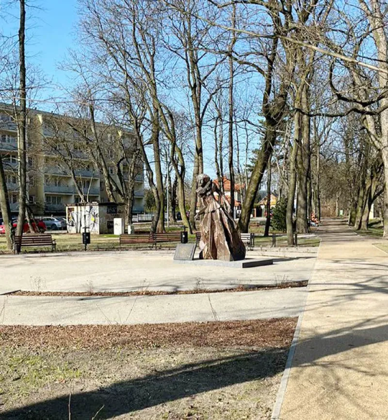 W parku widoczna jest rzeźba umieszczona na prostokątnej podstawie, otoczona przez nagą zieleń drzew i ławki, z alejkami prowadzącymi w różnych kierunkach.