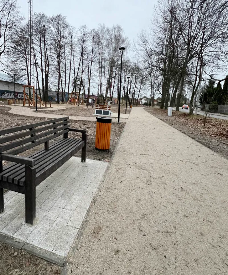 W parku znajduje się drewniana ławka oraz kosz na śmieci w kolorze pomarańczowym, a w tle są drzewa i plac zabaw. Ścieżka wzdłuż ławki jest wysypana drobnym żwirkiem.