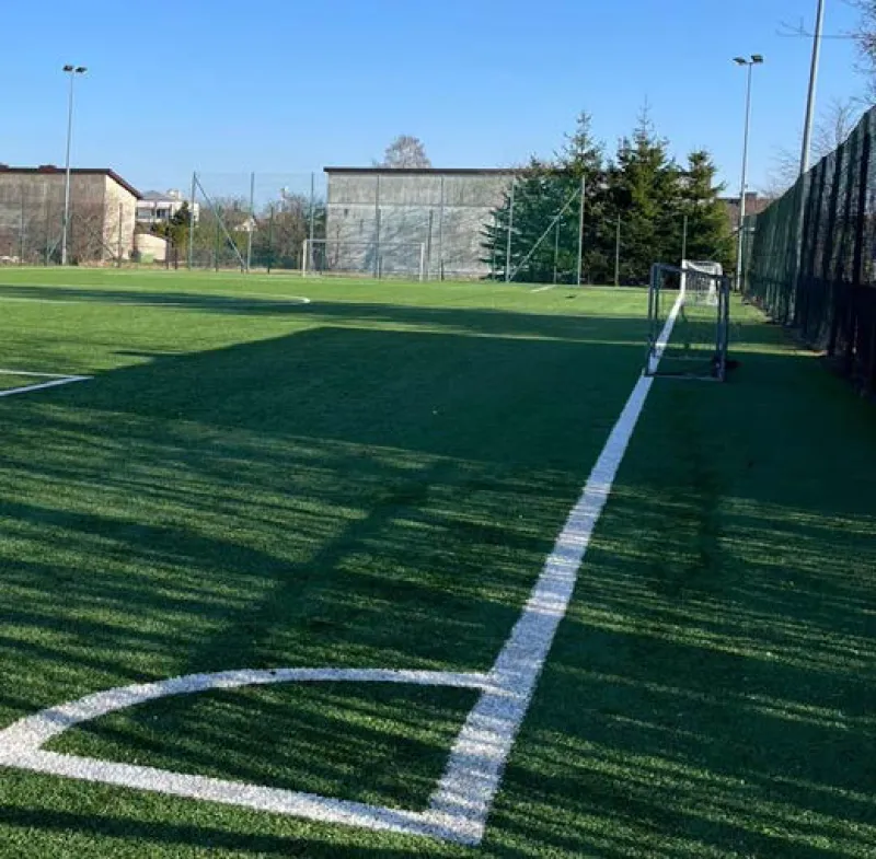 Pojedyncza bramka futbolowa stoi na sztucznej murawie boiska, w tle widać ogrodzenie oraz budynki.