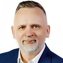 Portret mężczyzny w średnim wieku z krótkimi, jasnymi włosami i siwą brodą, ubrany w biały koszulę i granatową marynarkę.