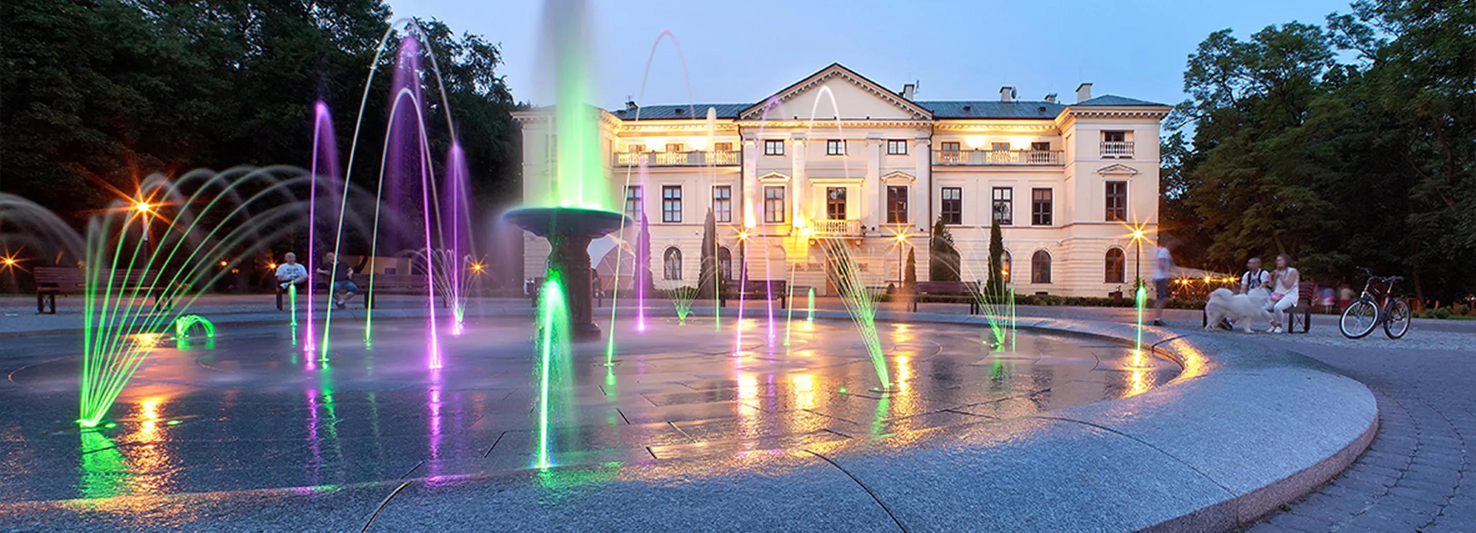 Kolorowa fontanna z podświetlanymi strumieniami wodnymi na tle oświetlonego budynku i ludzi siedzących na ławkach.