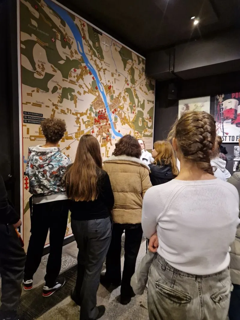 Grupa osób stoi plecami do kamery, wpatrując się w dużą mapę umieszczoną na ścianie, przedstawiającą teren z zaznaczonymi punktami, w pobliżu rzeki.