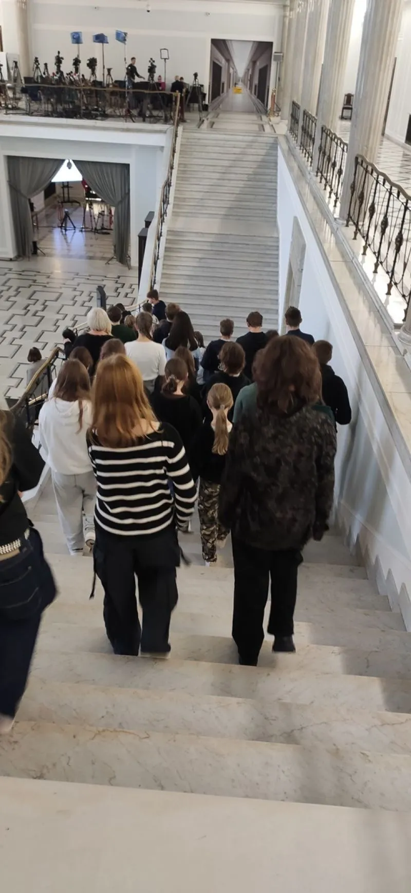 Grupa osób schodzi po marmurowych schodach w eleganckim wnętrzu budynku, w tle widoczne oświetlenie i sprzęt filmowy.