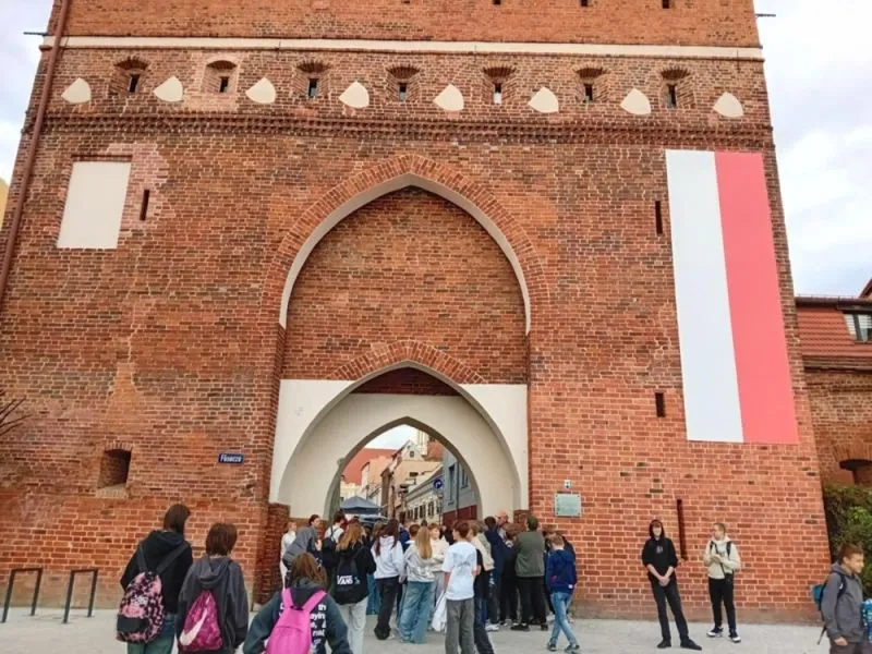 Grupa osób stoi przed dużą, czerwoną, ceglaną bramą z łukiem, na której wisi biało-czerwona flaga, w tle widoczne budynki.
