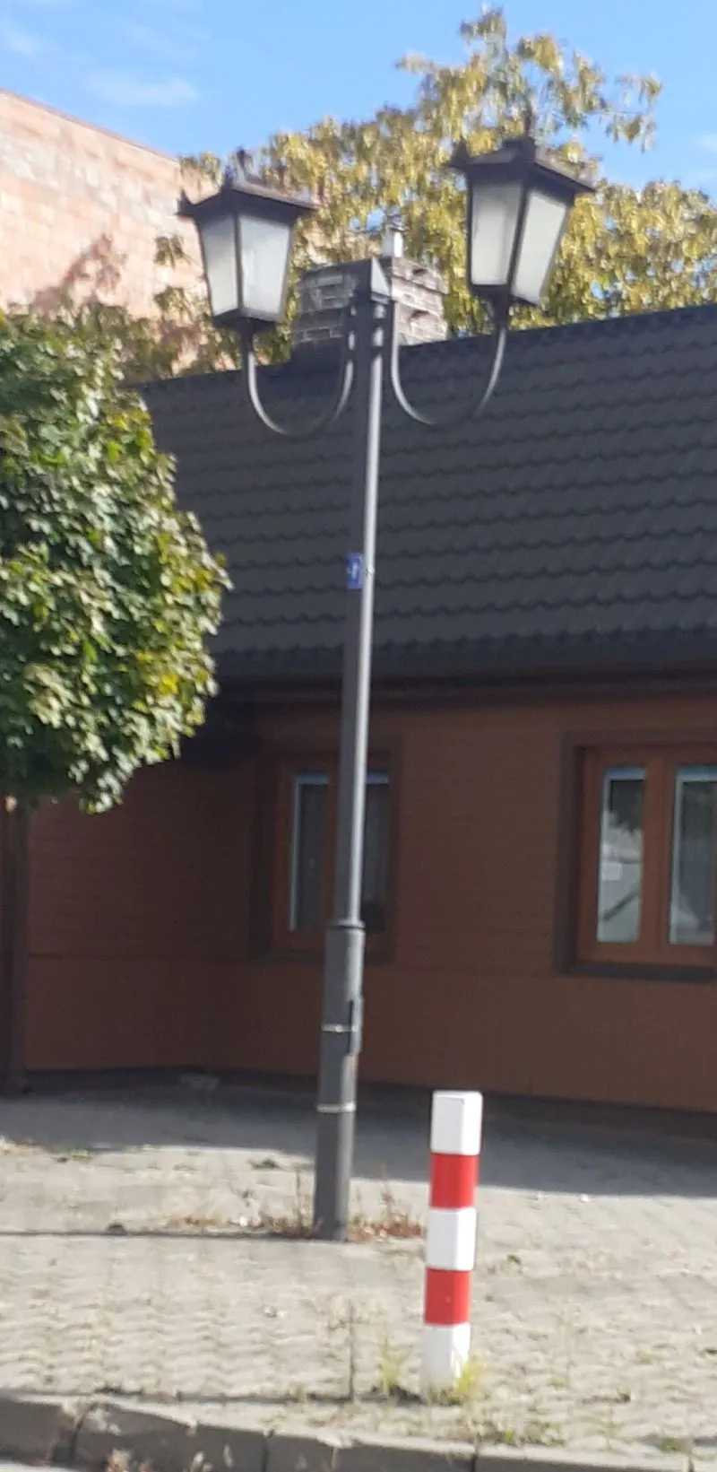 Latarnia uliczna stoi obok budynku z brązową elewacją i oknami w kolorze białym oraz piaskowym chodnikiem, na którym znajduje się słupek z czerwono-białymi pasami.