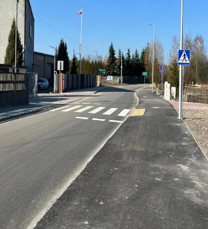 Ulica z przejściem dla pieszych, sygnalizacją i oznaczeniami drogowymi, otoczona drzewami i budynkami.