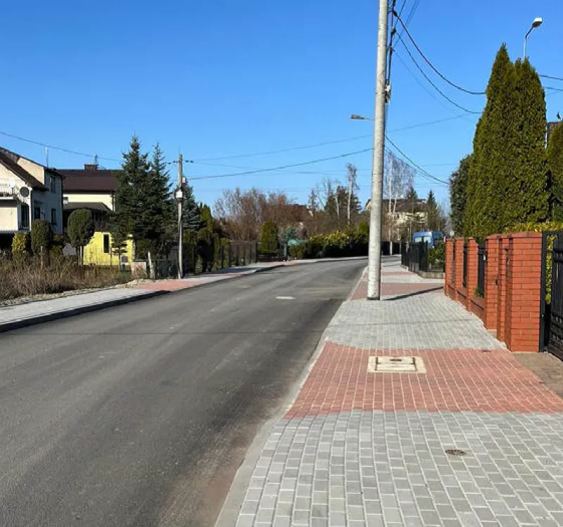 Ulica z chodnikiem, domami po lewej stronie oraz zielonymi drzewami i żywopłotem, i niebieskim niebem.