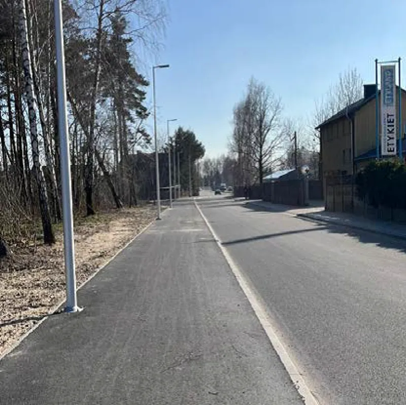 Na ulicy widoczne są latarnie, drzewka po lewej stronie oraz budynki po prawej, a w tle widać kontynuację drogi.